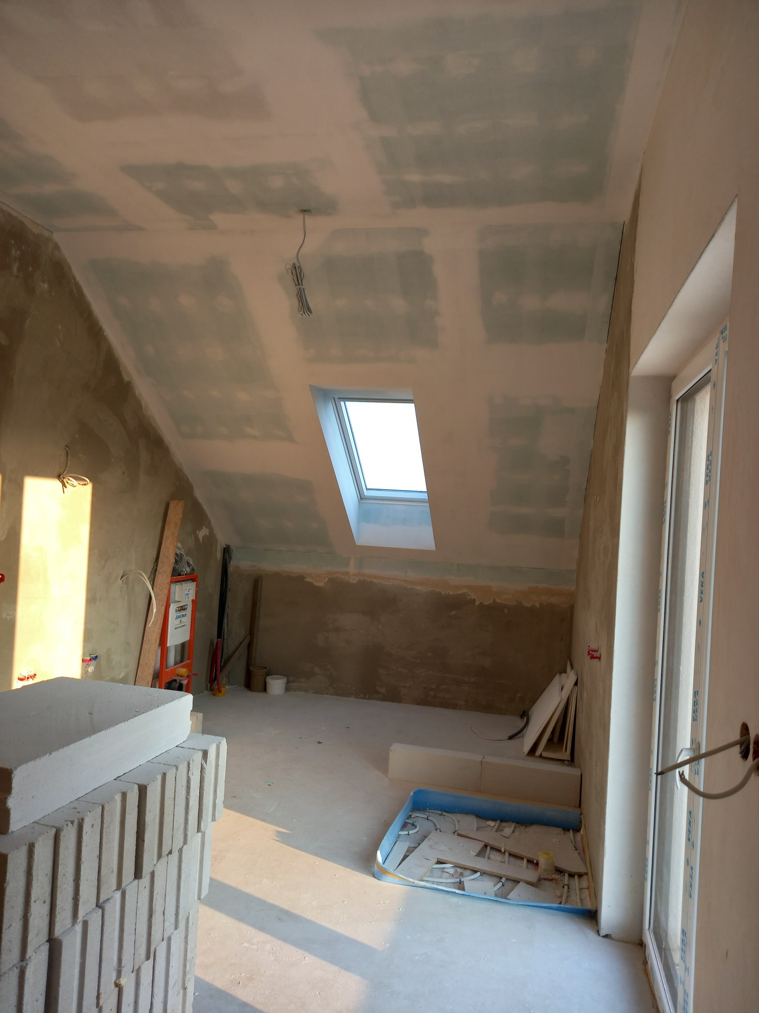 Poddasze w trakcie prac wykończeniowych: widoczne płyty kartonowo-gipsowe na skosach sufitu, okno dachowe, przygotowane ściany, materiały budowlane i narzędzia w pomieszczeniu.