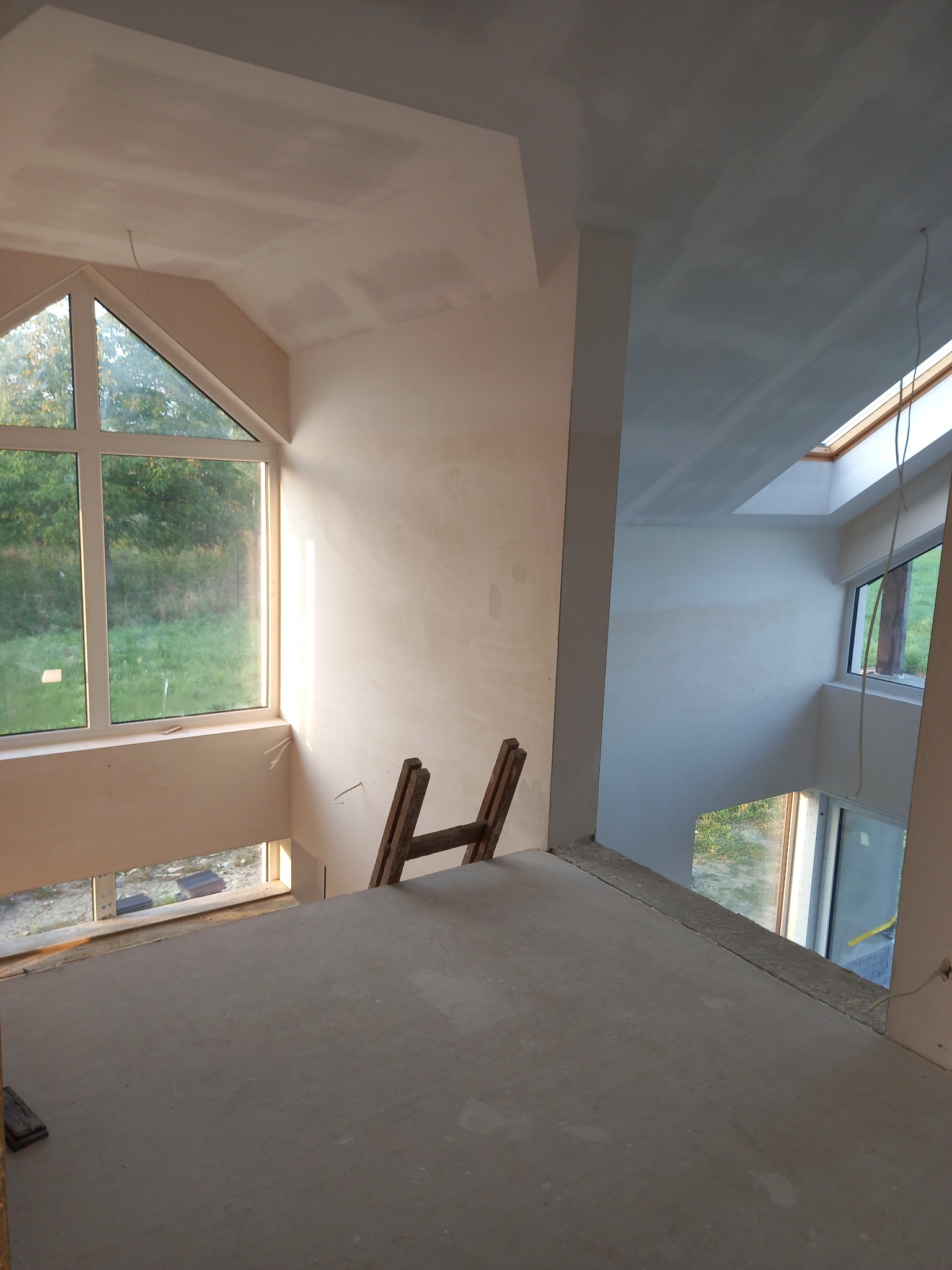 Wnętrze domu w trakcie prac wykończeniowych: ściany pokryte gładzią szpachlową, widoczne okno trójkątne, drewniana drabina oparta o ścianę, sufit z oknem dachowym, widok na zewnątrz przez okna.