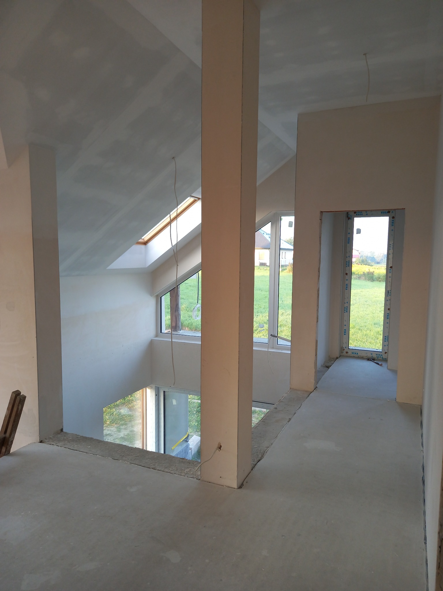 Wnętrze w trakcie remontu: surowe ściany, wnęki okienne, widoczne okno dachowe i pionowy słup konstrukcyjny, betonowa wylewka na podłodze, brak wykończenia, widoczne przewody elektryczne.