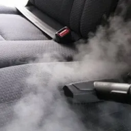 Czyszczenie parowe szarej tapicerki samochodowej z widocznym pasem bezpieczeństwa i klamrą; para unosząca się z dyszy urządzenia.