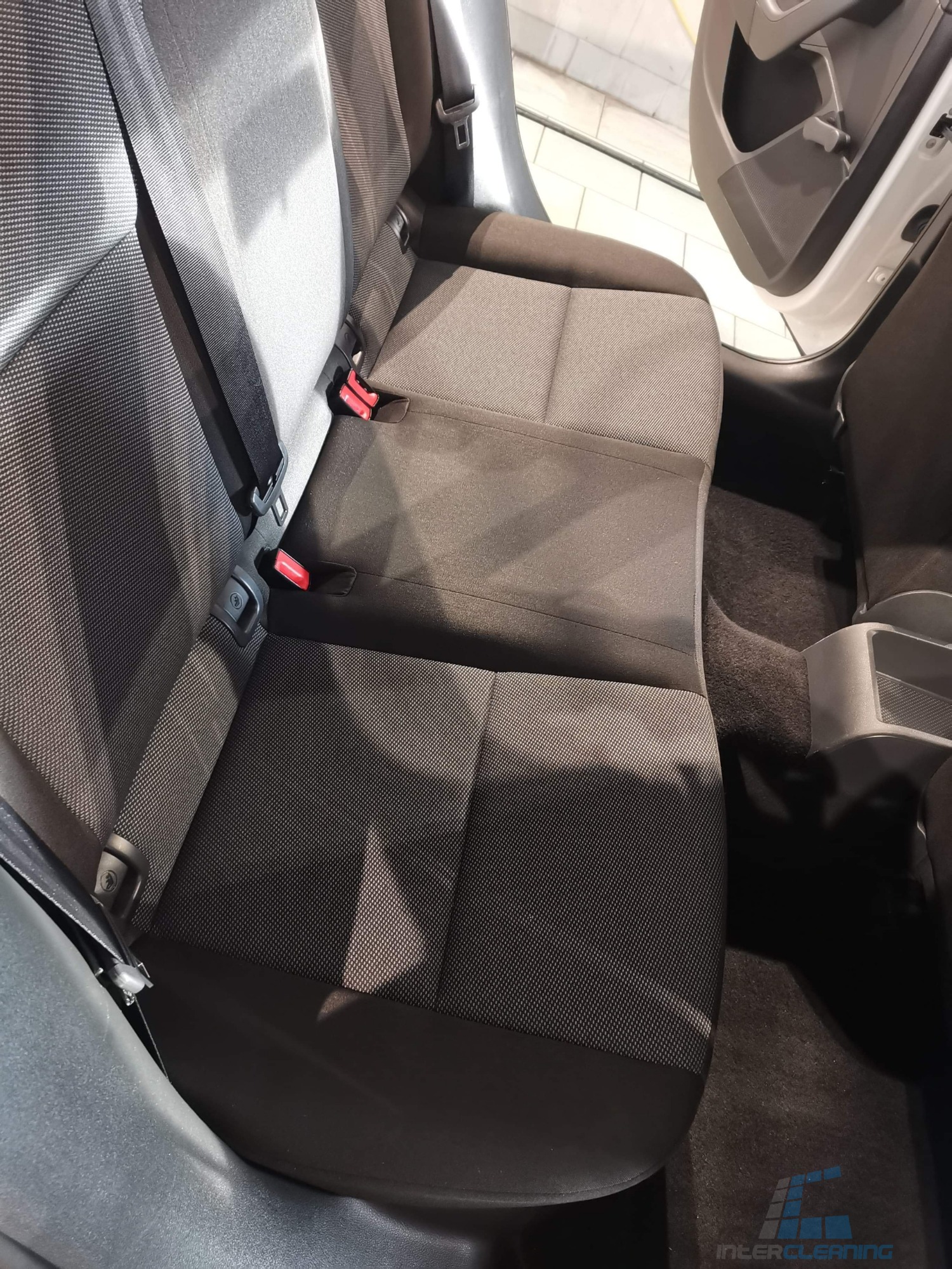 Wnętrze samochodu z widoczną tylną kanapą z szarej tapicerki materiałowej po praniu, z widocznymi zapięciami pasów bezpieczeństwa i fragmentem dywanika podłogowego.