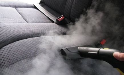 Czyszczenie parowe szarej tapicerki samochodowej z widocznym pasem bezpieczeństwa i klamrą; para unosząca się z dyszy urządzenia.