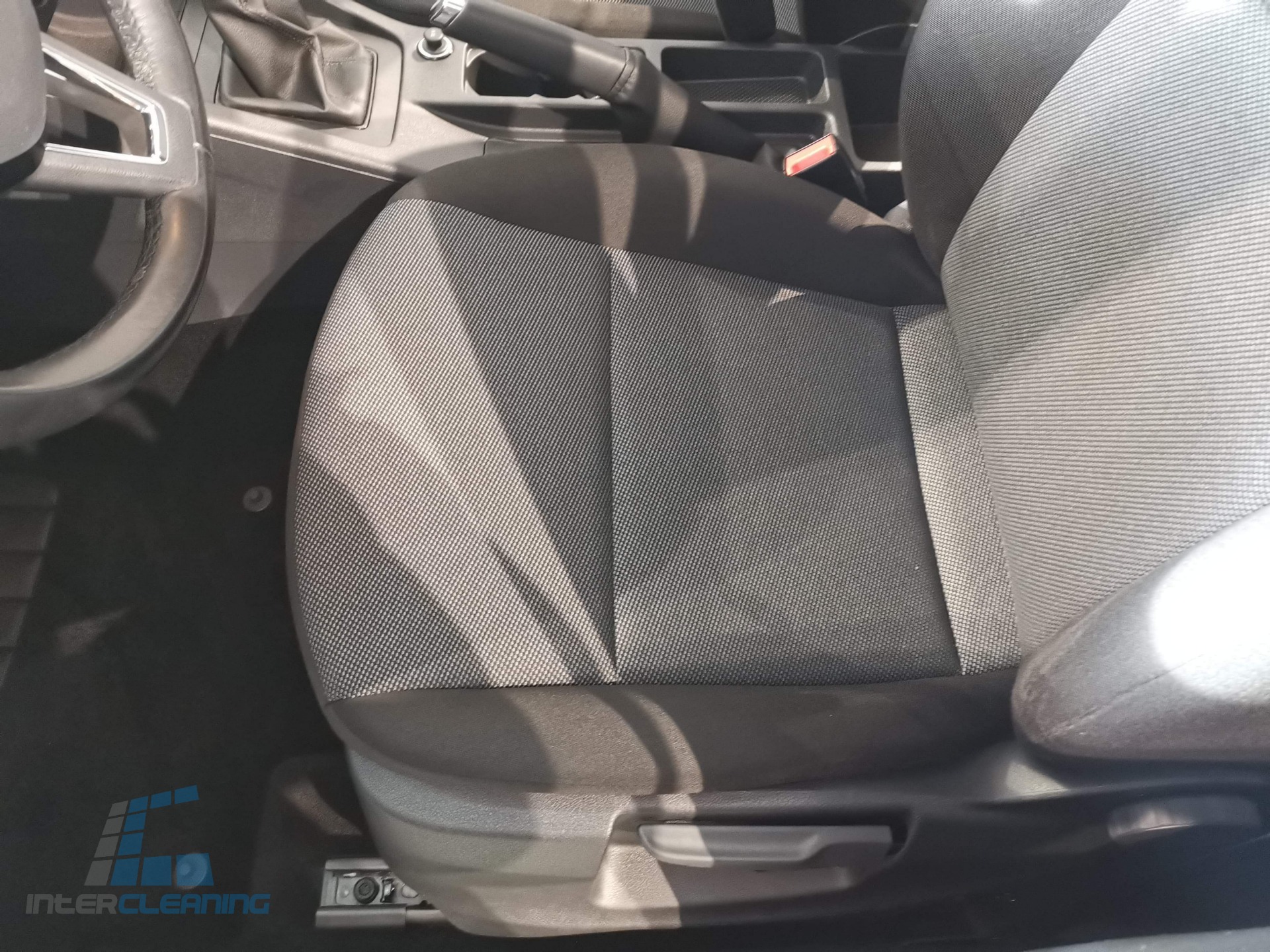 Wnętrze samochodu z wyczyszczonym, szarym fotelem kierowcy z tkaniny, widoczna kierownica i dźwignia hamulca ręcznego. W lewym dolnym rogu logo firmy sprzątającej.