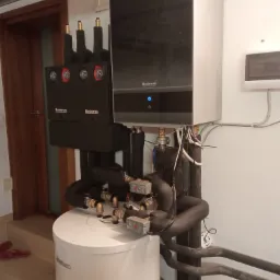 Kompletna instalacja kotła grzewczego Buderus z zasobnikiem ciepłej wody, widoczne rury i elementy przyłączeniowe, zamontowane w pomieszczeniu technicznym.