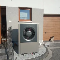 Zewnętrzna jednostka pompy ciepła Buderus, zainstalowana na kostce brukowej przed domem z drewnianą bramą garażową, w tle pies obserwujący otoczenie.