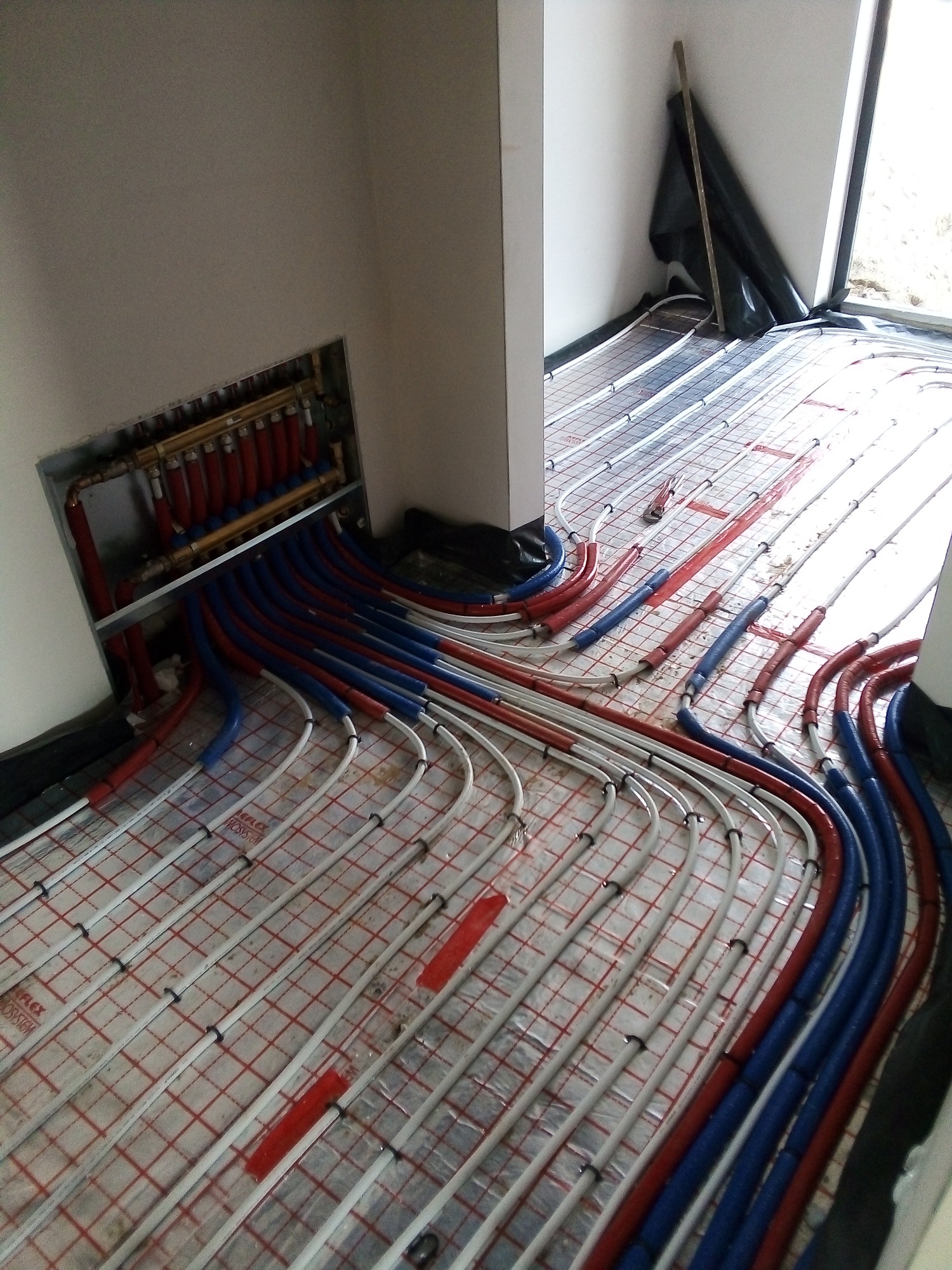 Rozłożone rury ogrzewania podłogowego na macie, z rozdzielaczem w ścianie, widok z góry pod kątem, częściowo zakryte folią ochronną.