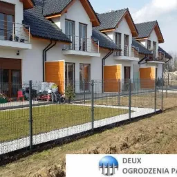 Szary panelowy system ogrodzeniowy otaczający posesję z nowymi domami w zabudowie szeregowej, widoczny biały żwir wzdłuż ogrodzenia.
