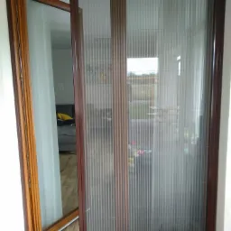 Rozłożona moskitiera plisowana w brązowej ramie, zamontowana w otwartych drzwiach balkonowych z drewnianą ramą, widok na wnętrze salonu.