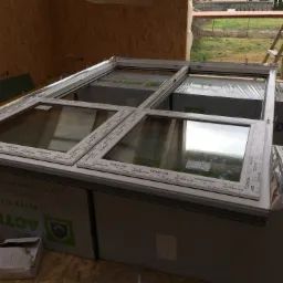 Nowe, jeszcze nie zamontowane okna z białymi ramami, oparte na opakowaniach styropianowych, w surowym wnętrzu budynku w trakcie budowy, drabina oparta o ścianę w tle.