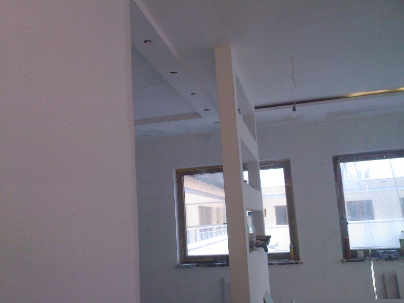 Świeżo pomalowane, jasne wnętrze z widoczną ścianą działową z otwartymi półkami, trzema oknami i wykończonym sufitem z punktowym oświetleniem.