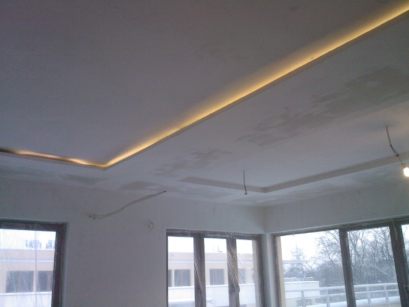 Pokój w trakcie remontu z widoczną konstrukcją pod sufit podwieszany z oświetleniem LED wzdłuż krawędzi, ściany w stanie surowym, duże okna z widokiem na zewnątrz.