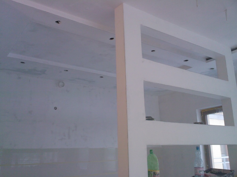 Wnętrze w trakcie prac wykończeniowych: widoczna biała ściana działowa z trzema prostokątnymi otworami, sufit z punktowymi lampami w trakcie montażu, ściany przygotowane do malowania, butelki...