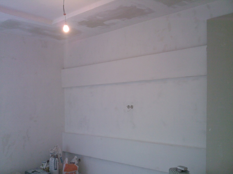 Wnętrze w trakcie prac wykończeniowych, ściany po szpachlowaniu z widocznymi wyprowadzeniami elektrycznymi, listwy dekoracyjne na ścianie, worki z zaprawą i narzędzia na podłodze, sufit...