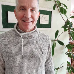 Uśmiechnięty mężczyzna z siwymi włosami w szarym swetrze z kapturem, stojący obok rośliny doniczkowej i obrazka z motywem ptaków w zielonej ramce.