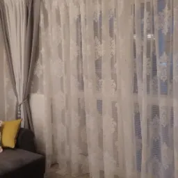 Salon z szarym narożnikiem i wzorzystymi, długimi zasłonami w oknie, część zasłon upięta sznurem.