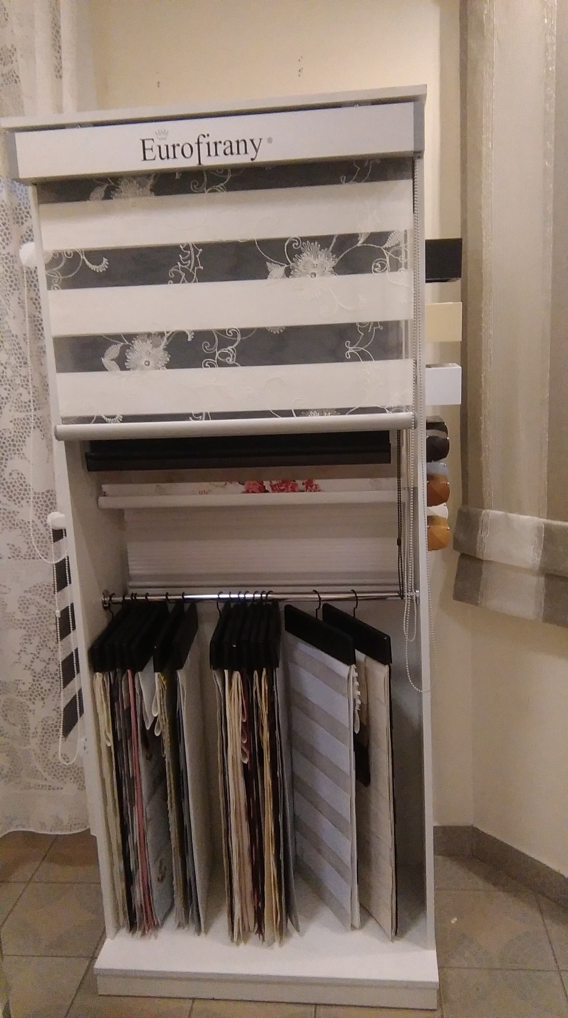 Ekspozycja różnych wzorów i kolorów rolet okiennych w salonie, z widocznym logo Eurofirany na górnej rolecie.