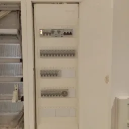 Otwarta szafka elektryczna z widocznymi bezpiecznikami i gniazdem, obok skrzynka z kablami, całość w jasnej tonacji.