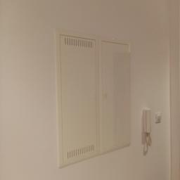 Info-Com - Biała skrzynka elektryczna z wentylacją na ścianie obok domofonu i włącznika światła.