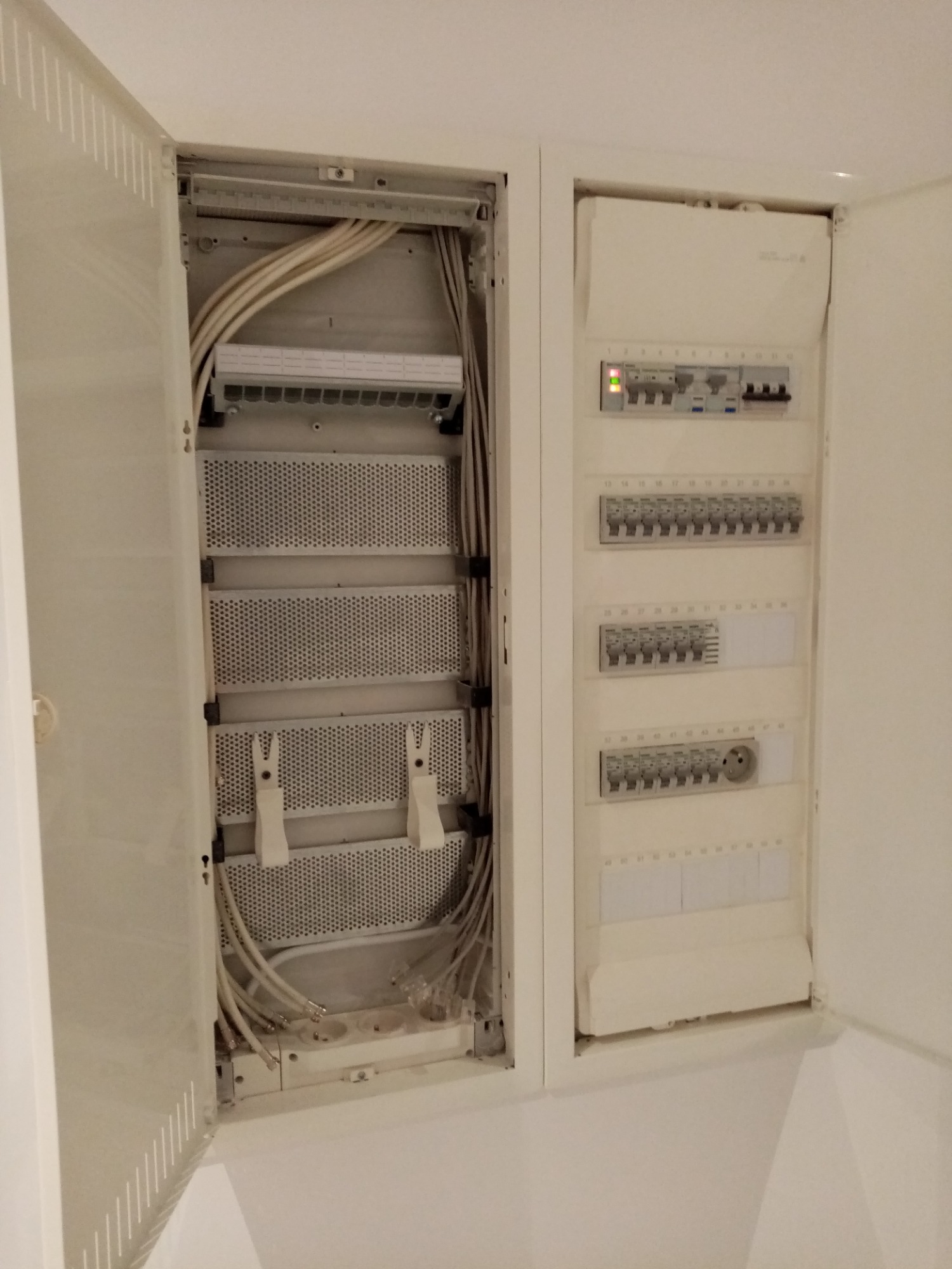 Otwarta, biała szafka rozdzielcza z widocznymi zabezpieczeniami, gniazdami elektrycznymi i kablami koncentrycznymi, zamontowana w ścianie.