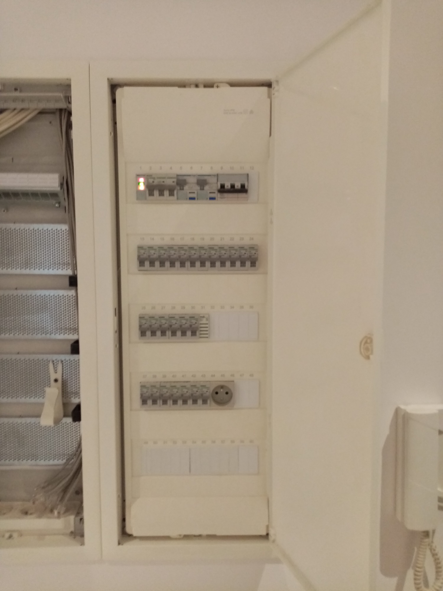 Otwarta szafka elektryczna z widocznymi bezpiecznikami i gniazdem, obok skrzynka z kablami, całość w jasnej tonacji.