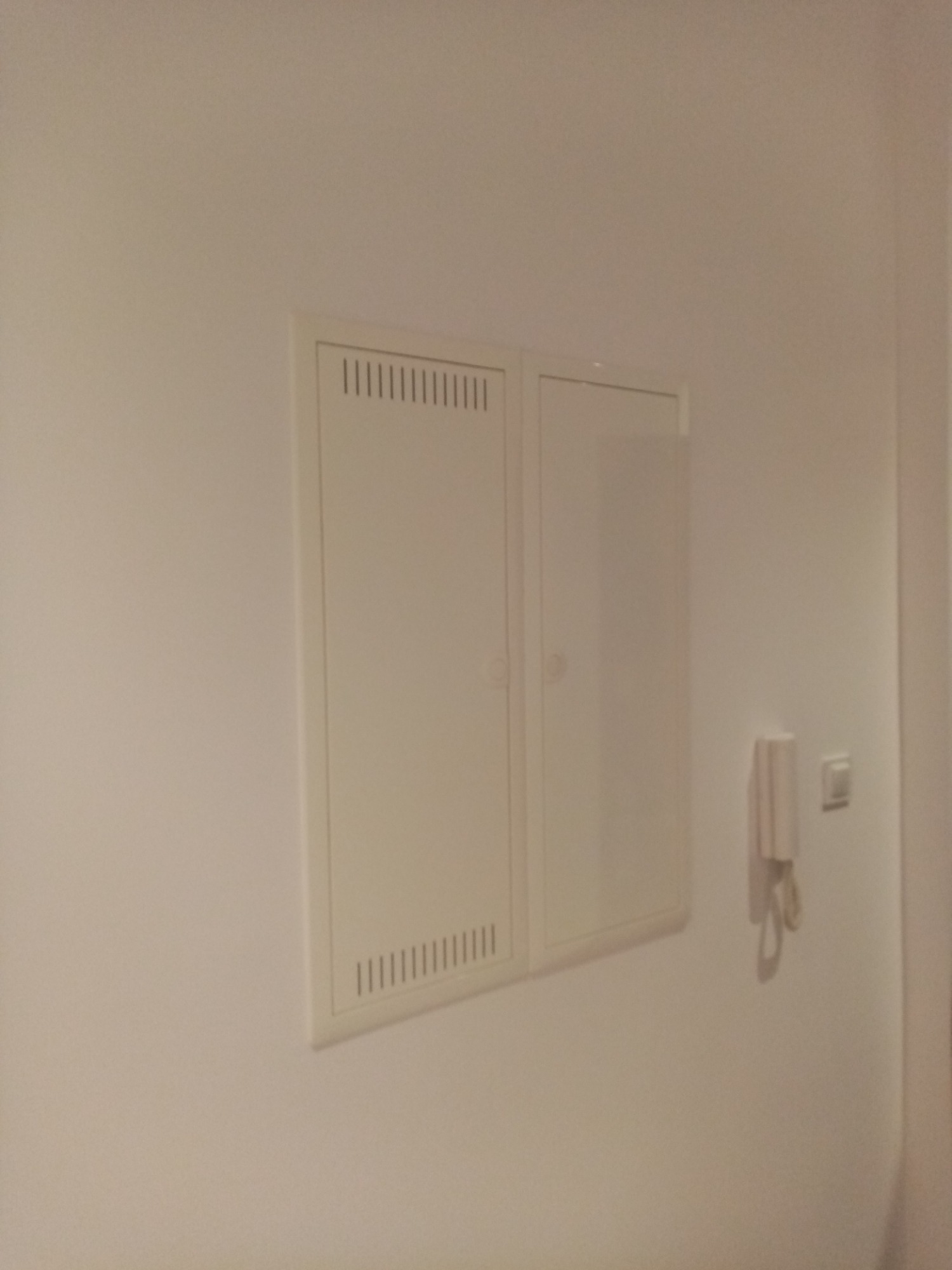 Biała skrzynka elektryczna z wentylacją na ścianie obok domofonu i włącznika światła.