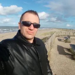 Autoportret mężczyzny w okularach i skórzanej kurtce na tle plaży z drewnianym płotem, ławkami i osobami w oddali, pod błękitnym niebem z chmurami.