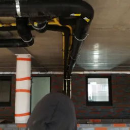 Czarne i żółte rury instalacyjne pod sufitem w surowym stanie, widoczne uchwyty mocujące, fragment ceglanej ściany z oknami, postać w bluzie z kapturem na pierwszym planie.