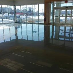 Błyszcząca, ciemna posadzka z widocznymi liniami podziału, odbijająca światło z dużych okien i szklanych drzwi wejściowych w nowym, jeszcze niewykończonym budynku.