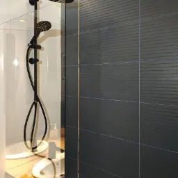 Nowoczesna kabina prysznicowa z czarną armaturą i ciemnymi płytkami z delikatnym wzorem, oddzielona szklaną ścianką, z widocznym blatem umywalki z jasnego drewna.