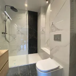 Nowoczesna łazienka z kabiną prysznicową typu walk-in, wykończona białymi i czarnymi płytkami imitującymi marmur, podwieszana toaleta i szafka z jasnego drewna.