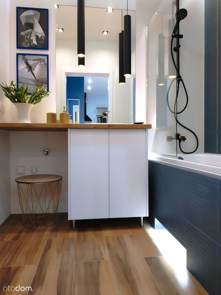Nowoczesna łazienka w Trzebini z drewnopodobną podłogą, białymi szafkami pod umywalką, lustrem z wiszącymi lampami i czarną armaturą prysznicową.