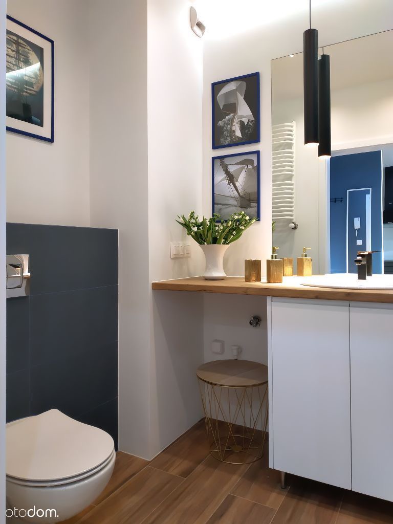 Nowoczesna łazienka z drewnianą podłogą, białymi meblami, kwiatami w wazonie i marynistycznymi obrazami w niebieskich ramkach.