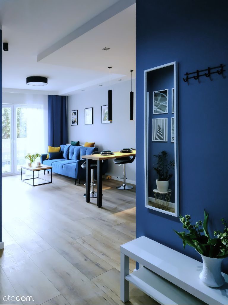 Nowoczesny salon z drewnopodobną podłogą, niebieską ścianą, minimalistycznym oświetleniem i błękitną sofą z żółtymi poduszkami, widziany z perspektywy wejścia.