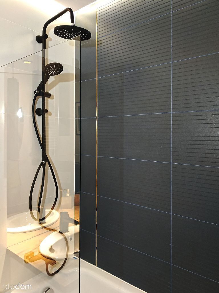 Nowoczesna kabina prysznicowa z czarną armaturą i ciemnymi płytkami z delikatnym wzorem, oddzielona szklaną ścianką, z widocznym blatem umywalki z jasnego drewna.