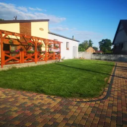 Zadbany trawnik przed domem z drewnianą pergolą zdobioną kwiatami w donicach, kolorowa kostka brukowa na podjeździe, w tle budynek w trakcie budowy.