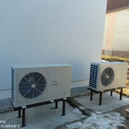 Dwie jednostki zewnętrzne pompy ciepła Galmet Prima R32 zamontowane na metalowych konstrukcjach, przed budynkiem z fragmentami śniegu na ziemi.