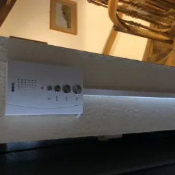 Biały panel domofonowy marki Eura zamontowany na teksturowanej ścianie wewnątrz domu z drewnianą konstrukcją stropu. Widoczne przyciski i otwory mikrofonu.