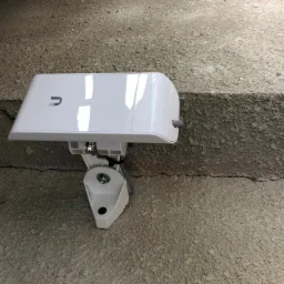 Biała antena sieci bezprzewodowej zamontowana na szorstkiej, betonowej ścianie z widocznym kablem. Antena posiada logo producenta w lewym górnym rogu.