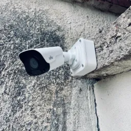 Biała kamera monitoringu UNV zamontowana na szarej, chropowatej ścianie zewnętrznej z fragmentami cegły.