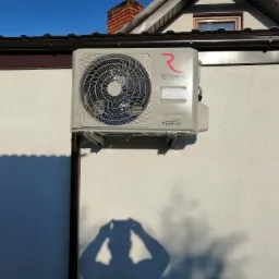 Zewnętrzna jednostka klimatyzacji marki Rotenso zamontowana na jasnej elewacji domu, widoczny cień osoby robiącej zdjęcie.