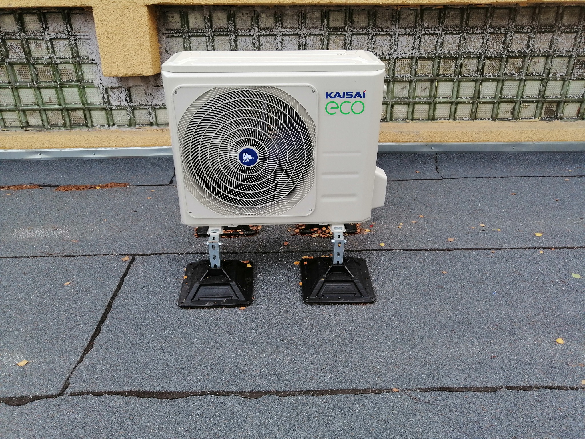 Biała jednostka zewnętrzna klimatyzacji Kaisai Eco zamontowana na dachu, widoczne czarne podstawy montażowe i fragment ściany z luksferami w tle.