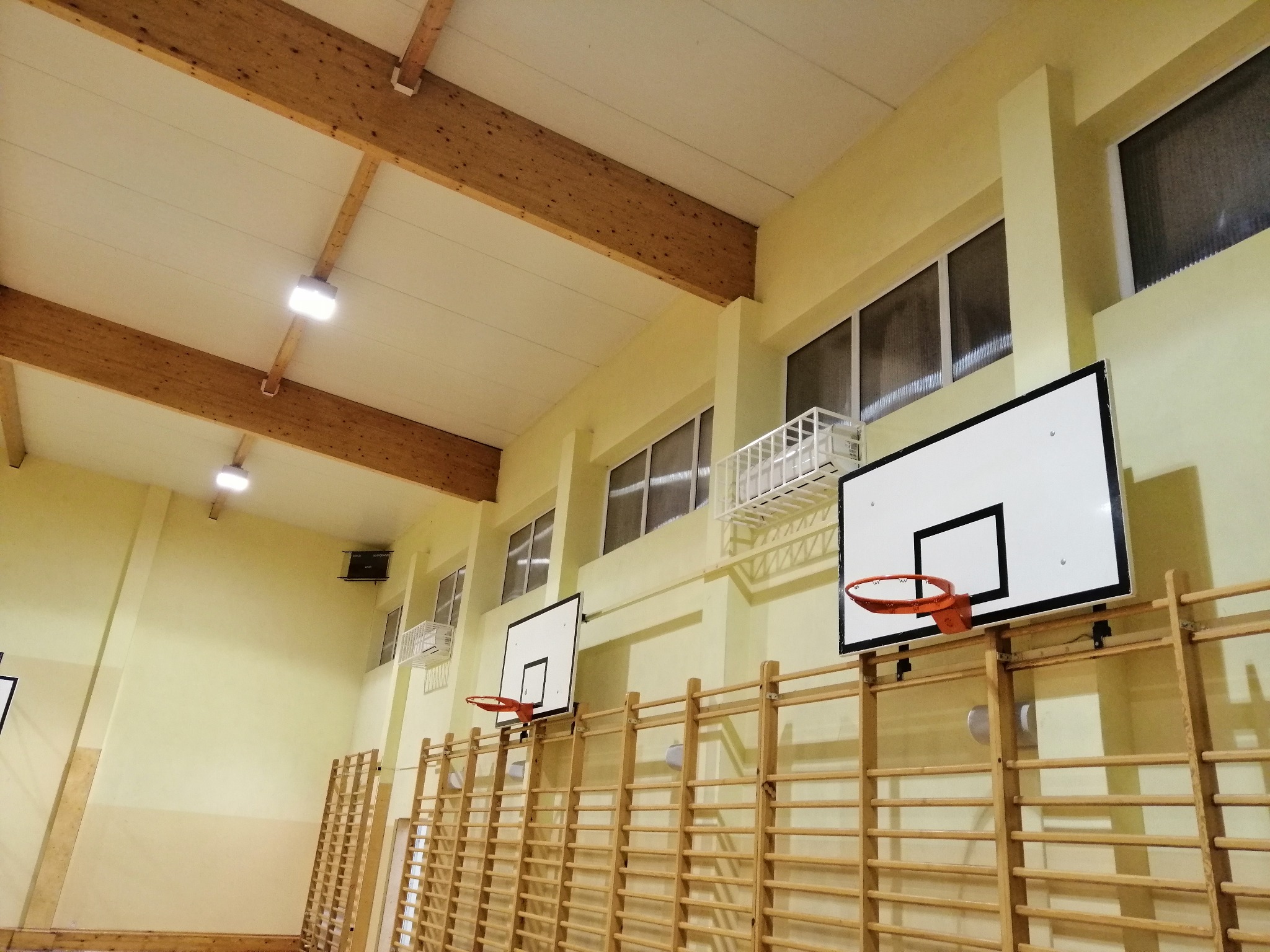 Wnętrze sali gimnastycznej z drewnianym sufitem, oświetleniem LED, oknami z kratkami i zamontowanymi klimatyzatorami oraz drabinkami gimnastycznymi z koszami do koszykówki.