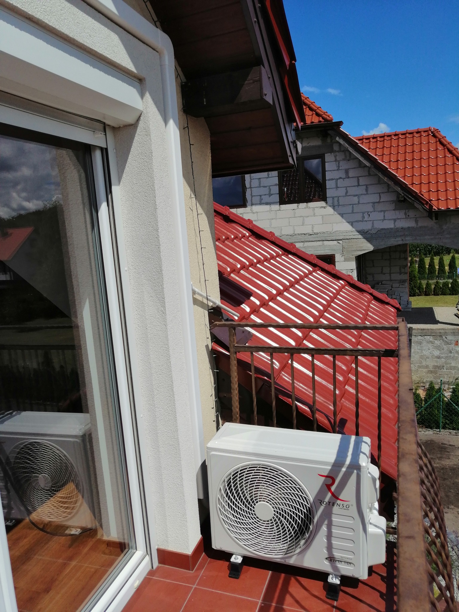 Biały klimatyzator Rotenso Inverter zamontowany na balkonie z czerwoną terakotą, widok z góry na czerwoną dachówkę domu w budowie i błękitne niebo.