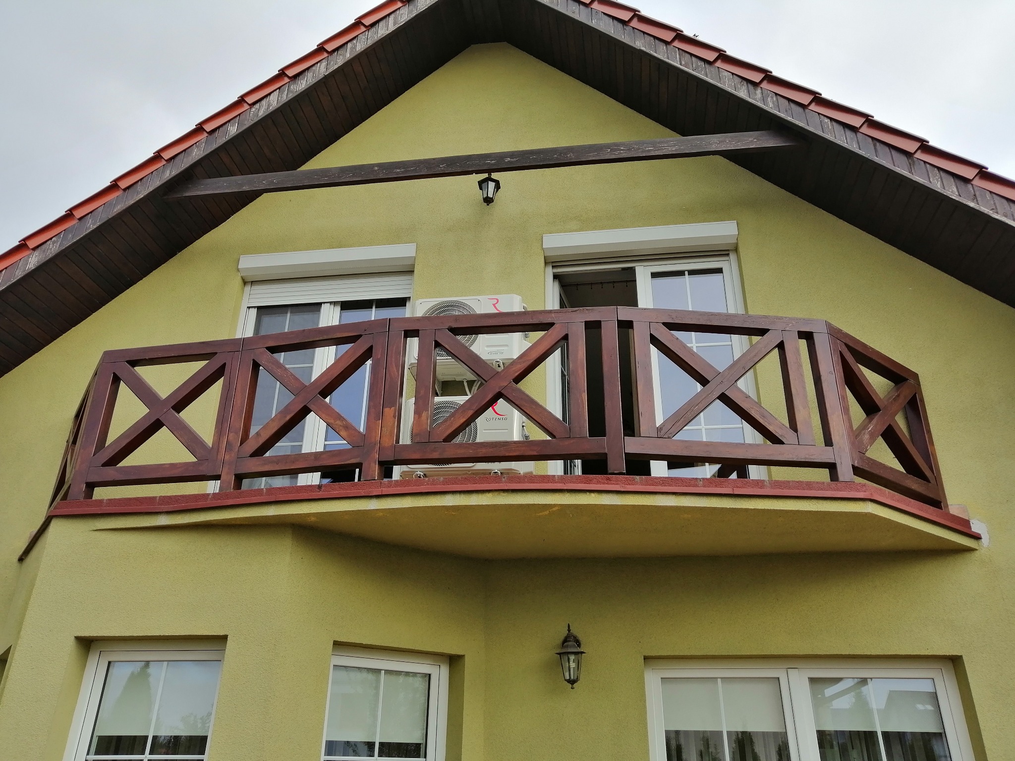 Zewnętrzna jednostka klimatyzacji Rotenso zamontowana na balkonie domu o żółtej elewacji, widoczna przez drewnianą balustradę z krzyżowym wzorem.