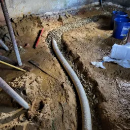 Wykop w gruncie z ułożoną perforowaną rurą drenażową obsypaną żwirem, widoczne narzędzia i wiaderka, surowy stan piwnicy w tle.