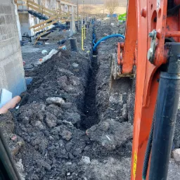 Pomarańczowa koparka w trakcie wykonywania wykopu pod rury na terenie budowy szeregowych domów, widoczne ułożone rury w wykopie oraz elementy konstrukcyjne budynków w tle.