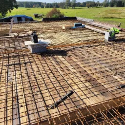 Siatka zbrojeniowa na deskowaniu szalunkowym, widok z góry na przygotowanie do wylewki, w tle krajobraz wiejski i błękitne niebo.