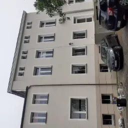 Jasna elewacja wielopiętrowego budynku z widocznymi oknami, klimatyzatorem i zaparkowanymi samochodami w pobliżu.