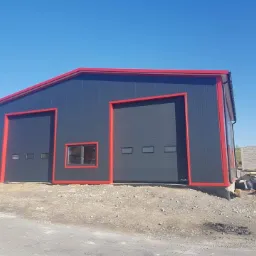 Nowoczesny, antracytowy budynek z blachy trapezowej z czerwonymi obramowaniami, wyposażony w dwie bramy segmentowe i okno, na placu budowy.