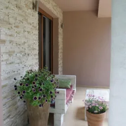 Wykończony balkon z dekoracyjną ścianą z imitacji kamienia, drewnianą ławką z poduszkami i donicami z kwiatami.